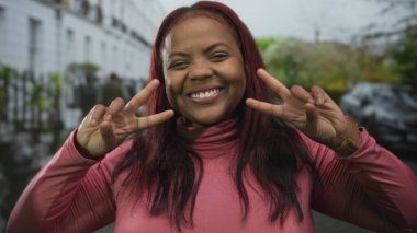 Pembe balıkçı yaka giyen kadın geniş bir gülümseme ve kentte barış işareti yapıyor; iyimserlik.