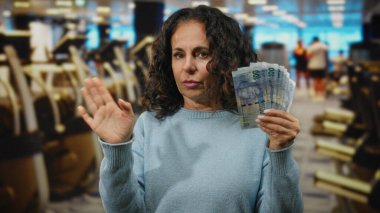 Orta yaşlı bir kadın, spor salonunda Güney Afrika Rand banknotları tutuyor. Egzersiz ekipmanları ve hareketli bir geçmişi olan, kapalı alandaki şüphelerini dile getiriyor..