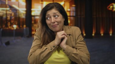 Latin kadın nazikçe gülümsüyor, elleri sımsıkı kapalı, gözleri kapalı, kahverengi ceketli, sarı gömlekli, sükunetli..