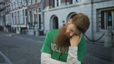 Yeşil gönüllü gömlekli ve beyaz uzun kollu fanilalı adam şehir caddesinde yumruk üzerinde dinleniyor; tefekkür.