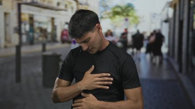 Güneşli, kalabalık bir caddede, dükkanlar ve yayalarla dolu, siyah tişörtlü bir adam karnını sıkıyor..