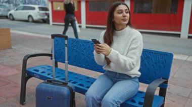Sokakta bir bankta oturan bir kadın mavi bavullu bir akıllı telefon kullanıyor, şehir içi bir seyahat sahnesini kaldırımda günlük kıyafetleriyle çekiyor..
