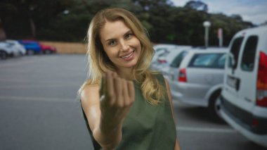 Parkta el kol hareketi yapan bir kadın, gülümseyen ve kamerayla meşgul olan, güneşli bir günde park etmiş arabalar ve yemyeşil ile çevrili..