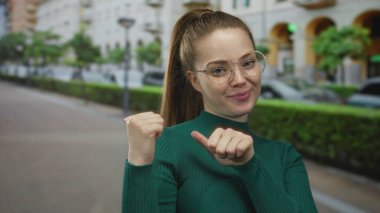 Yeşil kazak giyen ve parmaklarını kendisine gösteren kadın çitin ortasında kaldırım ve bina cephesi sokakta;.