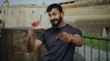 Roma 'da antik mimarinin önünde şakacı bir jest yapan sakallı İspanyol bir adam, tarihi bir taş arka plana işaret ediyor ve gülümsüyor..