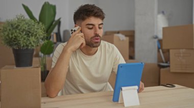 Hispanik adam akıllı telefonu kulağına tutuyor ve binadaki karton kutular arasında tablet ekranı tıklatıyor; hazırlık.