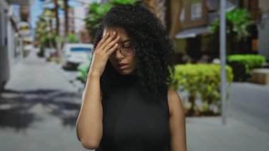 Kıvırcık saçlı ve gözlüklü genç İspanyol kadın canlı bir sokakta dikiliyor. Açık şehir ortamında, bulanık arka planda düşünceli görünüyor..