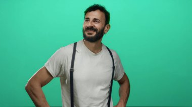 Sakallı İspanyol sakallı, elleri kalçasında canlı yeşil bir arka plana yaslanmış bir şekilde gülümseyen, stüdyo ortamında günlük beyaz gömlek ve pantolon askısı takan bir adam.