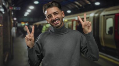 Gri balıkçı yaka kazaklı gülümseyen İspanyol adam tren istasyonunda iki eliyle iki barış işareti gösteriyor. İyimserlik..