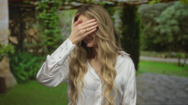 Parkta yeşil yapraklı bir kadın, açık beyaz gömlekli, yemyeşil ve doğal ortamda, yüzü elle kaplanmış bir şekilde duygusal tepki veriyor..