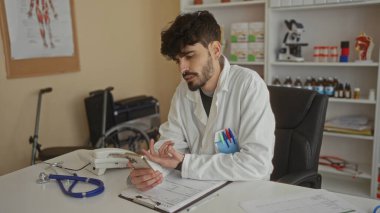 Klinikteki genç İspanyol bir adam video görüşmesi için akıllı telefonu kullanıyor. Üzerinde steteskop ve evrak gibi tıbbi ekipmanlar olan laboratuvar önlüğü var..