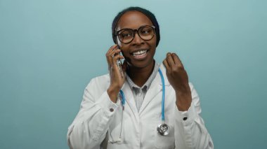 Akıllı telefondan konuşan bir kadın, boynunda steteskopla, izole edilmiş mavi bir arkaplan ile konuşuyor. Duygularını jestlerle ifade ediyor ve gözlük takıyor..