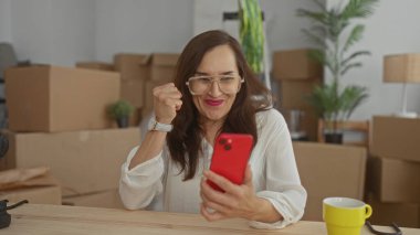 Yeni evde kırmızı telefon kullanan bir kadın taşınan kutularla çevrili. Ev içinde taşınma ve dijital teması vurguluyor.