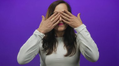 İspanyol kadın bir stüdyo ortamında mor bir duvara karşı iki eliyle gözlerini kapatır; sürpriz şaşkınlık merak.