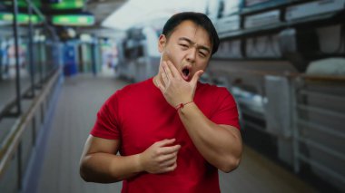 Bir yolcu gemisinde diş ağrısı çeken, acı dolu bir ifadeyle kırmızı tişört giyen genç bir adam, dış mekanın huzurlu arka planının ortasındaki rahatsızlığı yakalıyor..