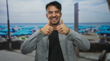 Mavi şemsiyeli ve arka planda sakin okyanus sularıyla pitoresk bir sahilde baş parmağıyla gülümseyen İspanyol genç adam..