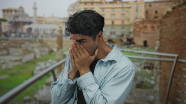 Genç adam, arkeolojik kazı bölgesindeki eski tuğla bina kalıntılarının yanında elleri ile yüzünü kapatıyor; endişe üzüntüsü.