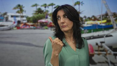 Orta yaşlı İspanyol kadın, arkasında yelkenli ve palmiye ağaçları olan bir limanı işaret ediyor..