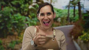 Açık hava parkındaki kahverengi önlüklü kadın, yeşil yeşilliklerle çevrili, parlak gülümseme ve doğal ortam ile pozitifliğini ifade eden, yaşam tarzı ve doğa temaları için uygun başparmak kaldırıyor..