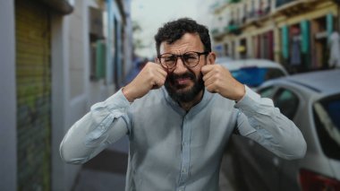 Sakallı İspanyol bir adam üzgün görünüyor ve arka planda arabalarla ve binalarla dolu bir sokakta ağlıyor..