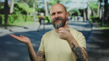 Şehir caddesinde sakallı, dövmeli, sarı gömlekli, açık havada güneş ışığı altında duran kel bir adam..