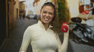 Genç İspanyol kadın şehir caddesinde kırmızı dambılı kaldırıyor enerjik bir şekilde dışarıda gülümsüyor sarı ceketli, hayat dolu pozitif bir atmosferde bulanık bir arka planı var.