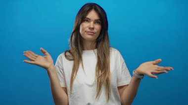 Genç İspanyol bir kadın, izole edilmiş mavi bir duvara karşı gelişigüzel alkışlıyor, beyaz bir gömlek giyiyor, rahat bir stüdyo ortamında dostça bir ifade sergiliyor..