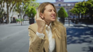 Güneşli bir şehir caddesinde, ağaçlar ve binalarla çevrili bulanık bir şehir arka planının önünde duygularını ifade eden bir kadın..