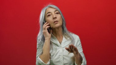 Beyaz saçlı yaşlı bir kadın akıllı bir telefonla konuşuyor, canlı kırmızı bir arkaplan karşısında duruyor ve anlamlı el hareketleriyle işaret ediyor..