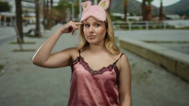 Kumsalda gece elbisesi giymiş genç sarışın bir kadın gündüz eğlencesine pembe uyku maskesi takmış komik suratlar yapıyor..