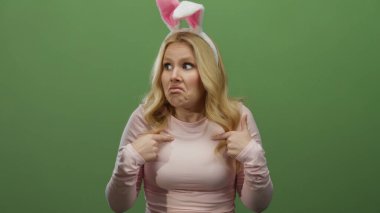 Sarı saçlı, tavşan kulağı takan ve pembe gömlekli bir kadın canlı yeşil bir arka plana esprili bir şekilde karşı çıkıyor..