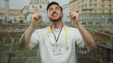 Doktor önlüğü ve steteskop giymiş sakallı genç adam, gülümsüyor ve Roma harabelerinin önünde yukarı doğru işaret ediyor..