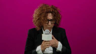 Gözlüklü İspanyol kadın mikrofon kullanıyor, canlı pembe bir arka plana karşı duruyor, siyah takım elbise giymiş, odaklanmış, ciddi bir muhabir rolünü somutlaştırıyor..