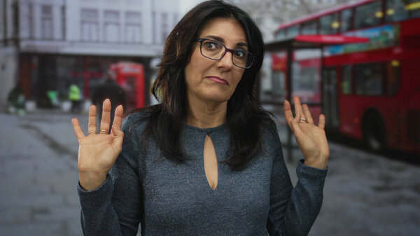 Middle aged hispanic woman wearing glasses and a blue sweater holding up hands in busy urban street; innocence.