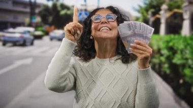 Orta yaşlı bir kadın elinde Endonezya rupia banknotları tutarken gülümsüyor ve kalabalık bir şehir caddesinde yukarı doğru işaret ediyor..