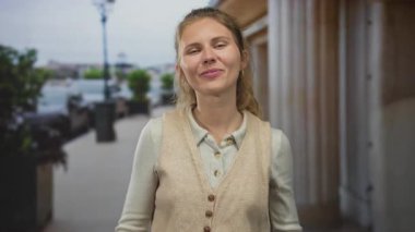 Genç bir kadın, bir caddeyi işaret ederken parmaklarını gösteriyor. Sarışın saçlar, parlak bir gülümsemeyle ışığa yakalanıyor ve şehrin önündeki bir binanın yakınındaki kameraya doğru gülüyor..