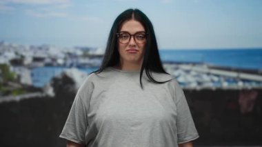 Gözlüklü kadın deniz kenarındaki limanın yanında duruyor. Kıvrımlı moda ve plaj sahnesini kucaklıyor. Rahat bir tavırla..