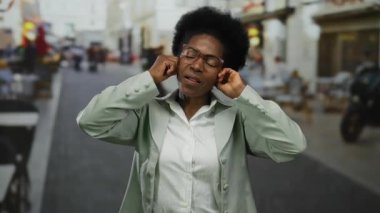 Gözlüklü Afrikalı Amerikalı bir kadın neşeli bir jest yapıyor. Kulaklarını kapatıyor ve canlı bir şehir caddesinde dilini çıkarıyor..