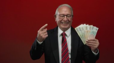 İş kıyafeti giymiş kıdemli bir adam, elinde Czech Koruna banknotları ile izole edilmiş kırmızı bir zemin üzerinde başarı ve zenginliği ima eden kendine güvenen bir ifadeyle..