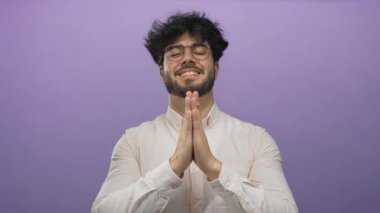 Beyaz gömlekli, sakallı ve gözlüklü genç adam mor arka planda el ele dua ediyor..