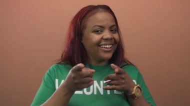 Yeşil tişört giyen, gülümseyen ve şeftali rengi stüdyo ortamında kameraya işaret eden gönüllü bir kadın..