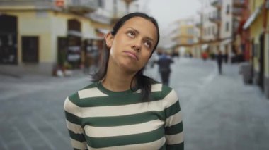 Şehirde hareketli bir sokakta düşünceli görünen genç bir kadın ifadeleriyle duygularını sergiliyor, açık havada çizgili bir gömlek giyiyor..