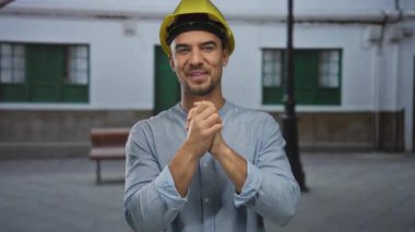 Açık havada kasketli genç İspanyol bir adam yeşil pencere panjuru ve sokak lambasına karşı rahat bir şekilde gülümsüyor..