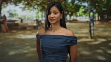 Bir kadın açık hava parkında dikiliyor. Düşünceli bir yüz ifadesi sergiliyor. Arka plandaki ağaçların arasından süzülen güneş ışığı, sakin bir atmosfer yaratıyor..