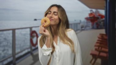 Gemi güvertesinde eli donut tutan kadın tırabzanın yanında gülümsüyor, gözleri kapalı; zevk düşkünlüğü.