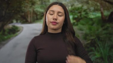 Ayakta duran kadın, elleri yeşil orman kıvrımlı yolda sık ağaçlar arasında mola veriyor; sakin duraklama.