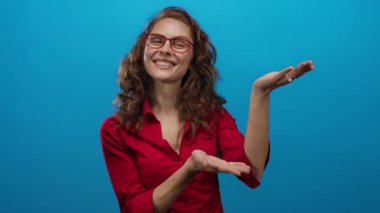 Kırmızı gömlekli kadın, mavi arka planda sunum yapar gibi ellerini uzatıyor, kıvırcık saçlar ve gözlüklerle gülümsüyor, izole ortamda pozitiflik tasvir ediyor..