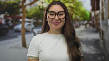 Gözlük takan ve günlük beyaz bluz giyen genç kadın, güneşli şehir caddesinde ağaçlarla kaplı ve trafiği bulanıklaştıran bir jest gösteriyor; iyimserlik.