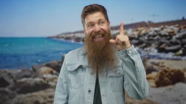 Kot ceket giyen, gülümseyen ve parmakla işaret eden bir adam güneşli kayalık bir sahilde, turkuaz deniz kenarında; iyimserlik.