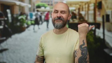 Şehir sokaklarında sakallı, dövmeli sıradan kıyafetler giyen bir adam açık şehir ortamında kendinden emin ve odaklanmış görünüyor..